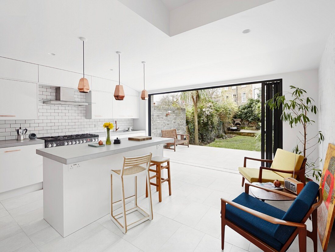 Moderne weiße Wohnküche mit freistehender Theke und Retro Sesseln mit Tisch, im Hintergrund offenes Schiebeelement mit Gartenblick