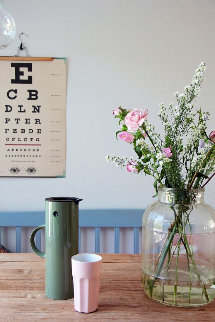 Flowers in large glass jar, thermos flask and beaker on wooden table with eye chart on wall in background
