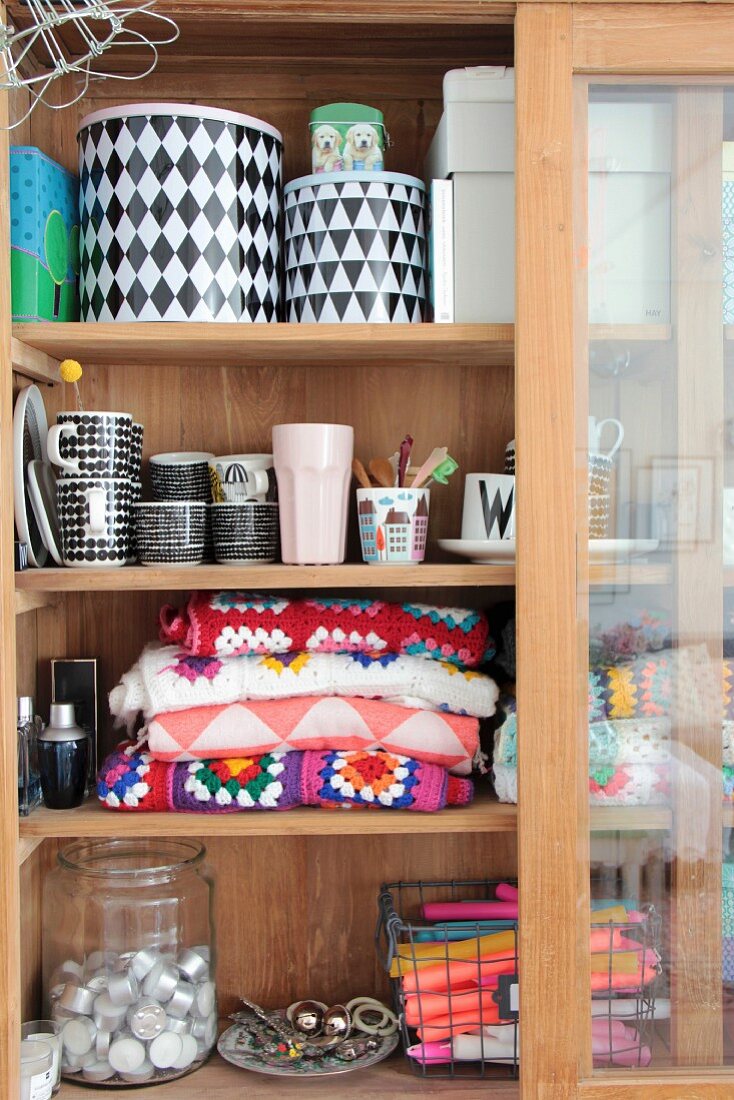 Colourful crocheted blanket, black and white crockery and candles in glass-fronted cabinet with sliding door