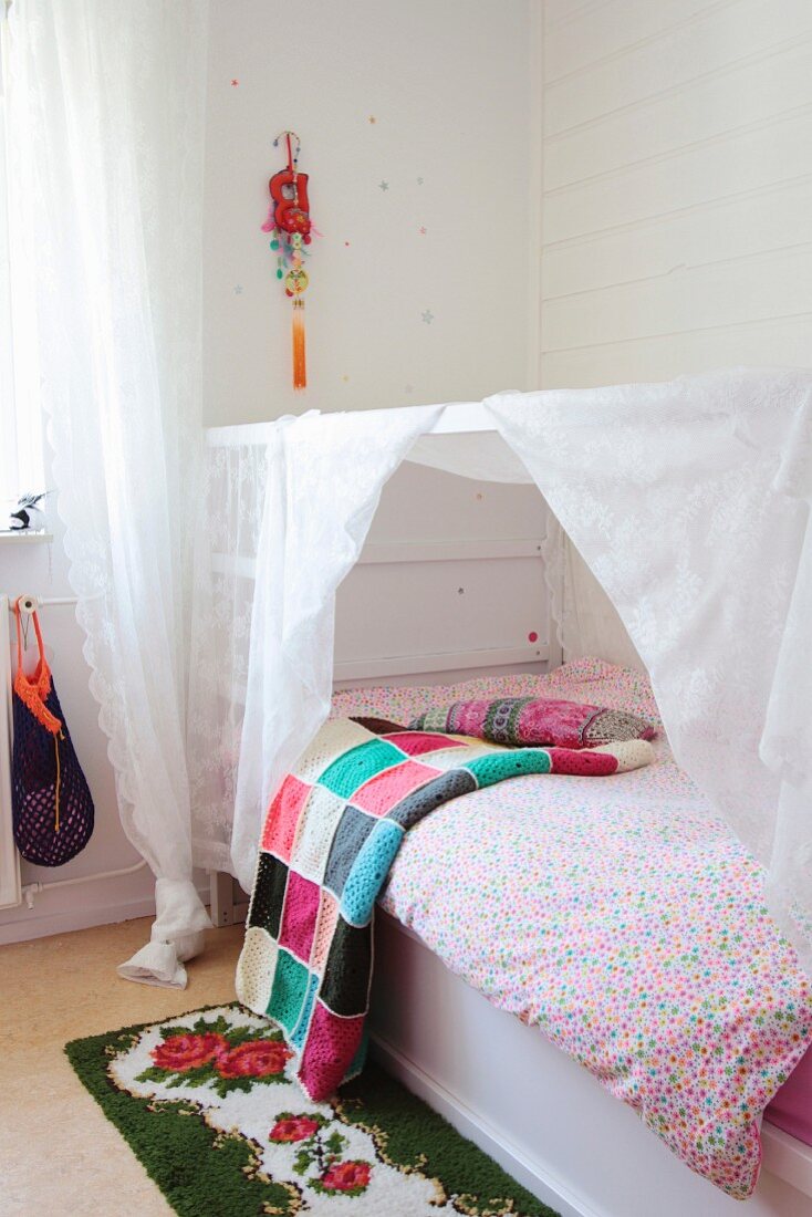 Colourful crocheted blanket, retro rose-patterned rug and child's white bed with canopy