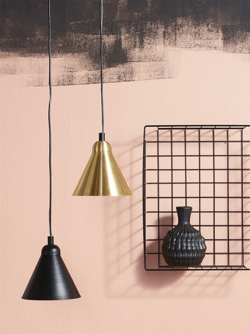 Two pendant lamps, one with a brass lampshade, one with a black lampshade, next to vase in wire basket mounted on wall painted pink and smudgy black