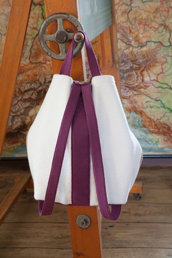 Selbstgenähte Rucksacktasche aus weißem Walkstoff mit violetten Trägern an Kurbel von Tafelgestell aufgehängt