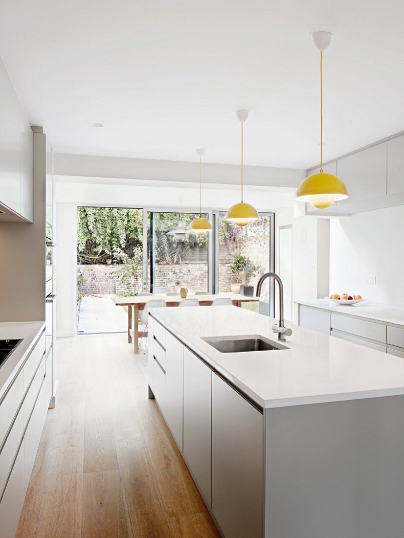 Gelbe Pendelleuchten in Reihe über weisser Kücheninsel in moderner Küche vor geöffneter Terrassentür