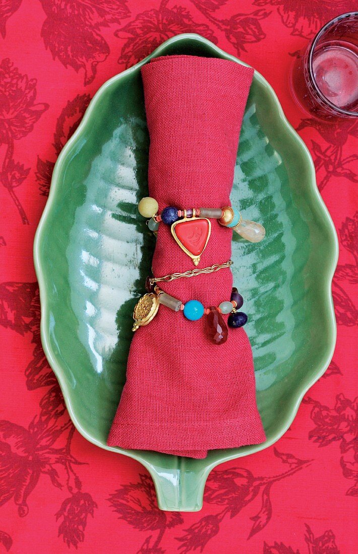 Red linen napkin tied with beaded chain on green leaf-shaped dish on red patterned tablecoth