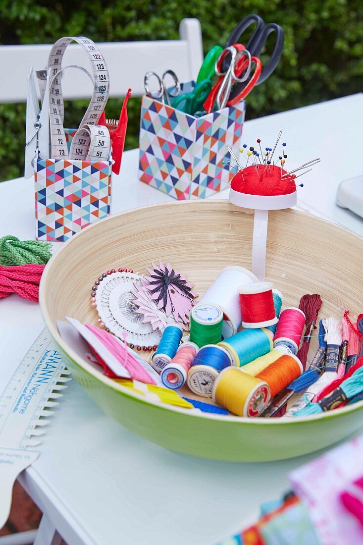Various sewing utensils (yarn, ties, pins, scissors) in a bowl and boxes on a table outdoors