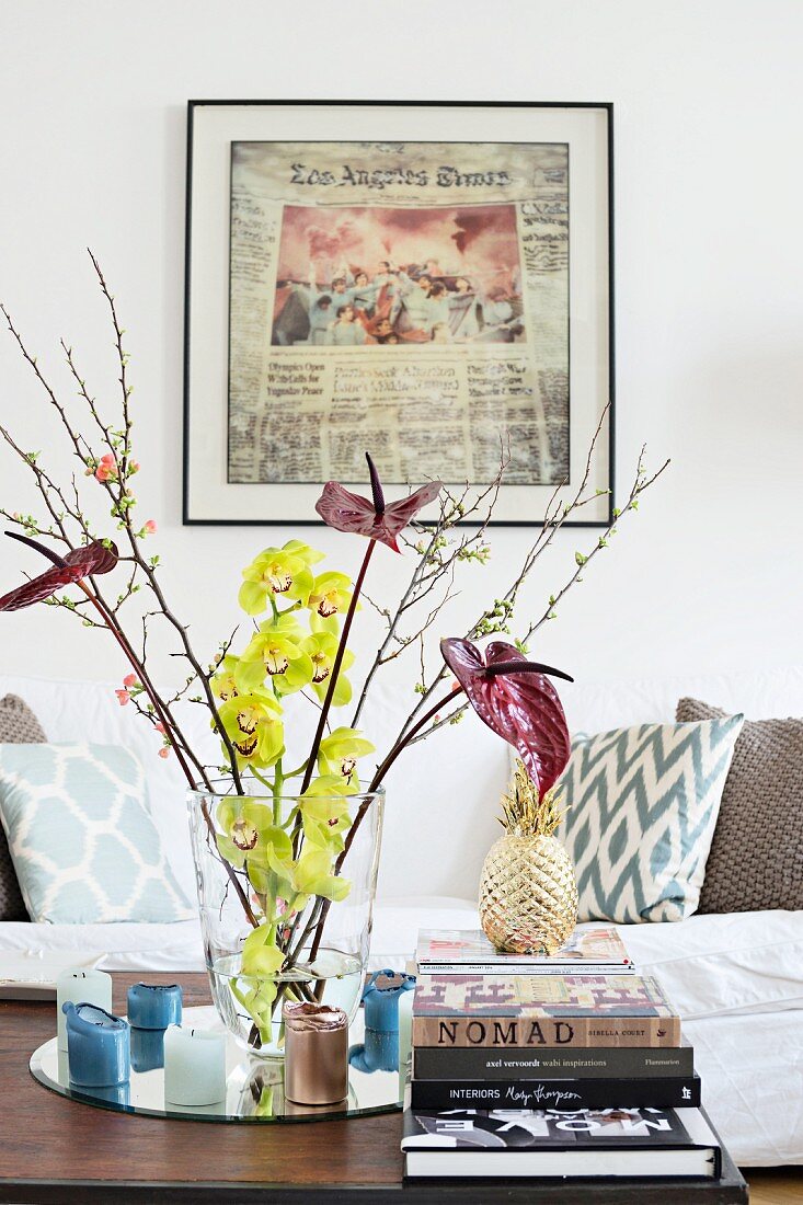 Glasvase mit Blüten, Kerzen und Bücherstapel vor gerahmtem Zeitungsausschnitt und Couch