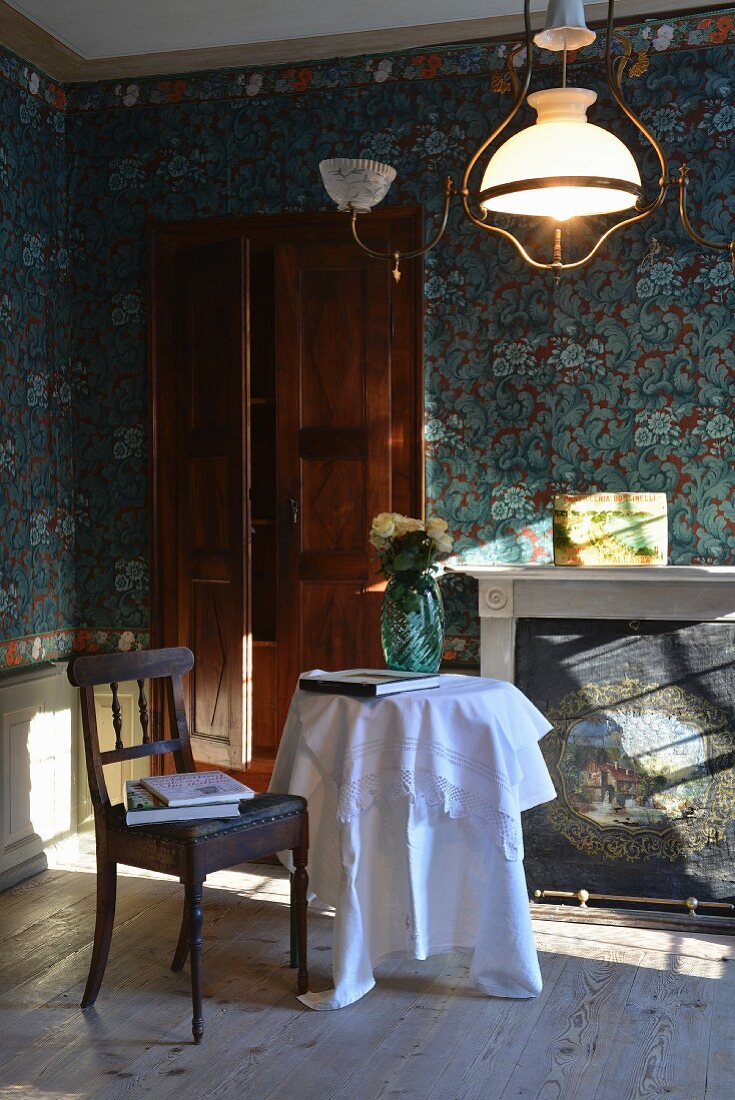 Vintage Holzstuhl mit Lederbezug und runder Tisch mit weisser Tischdecke vor Kamin, im Zimmer mit blau gemusterter Tapete