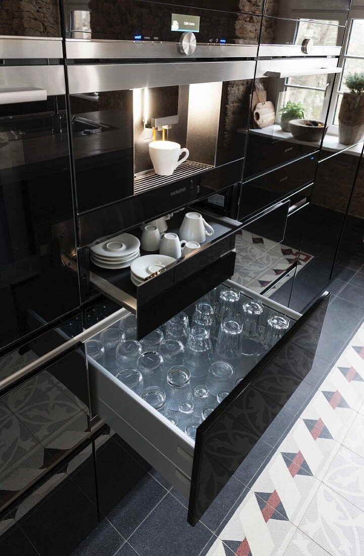 A kitchen cupboard with a shiny black surface, a coffee machine above an open drawer of cups and glasses
