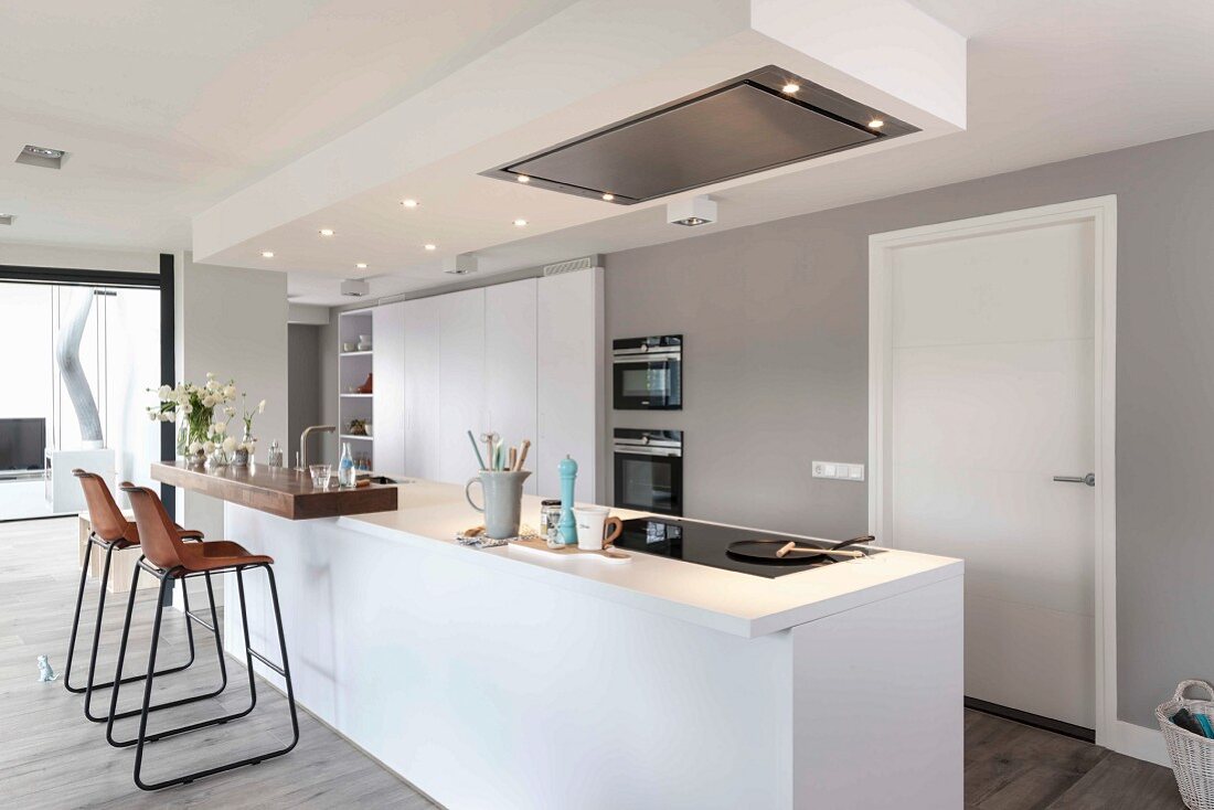 A modern white kitchen with an island with an illuminated, suspended ceiling panel and a extractor hood