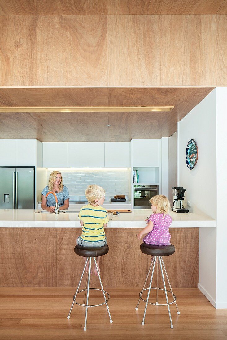 Kinder auf Barhockern und Mutter hinter freistehender Theke in moderner Küche mit abgehängter Holzdecke