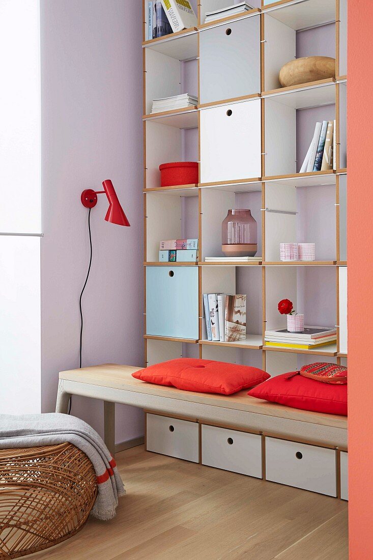 Floor-to-ceiling shelf with pastel-coloured flaps, open compartments and drawers with a bench in front of it with red cushions
