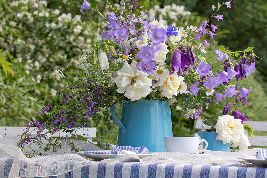Blau-weißer Blumenstrauß in einer Emaillekanne auf dem Gartentisch