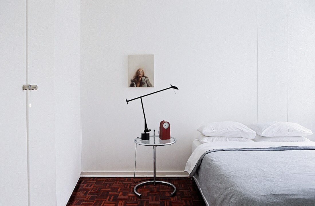 Alarm clock and table lamp on glass table next to double bed in minimalist, white guest room
