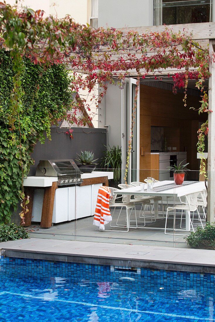 Pool mit blauen Mosaikfliesen und Terrasse mit Outdoor-Küche hinter Glasbrüstung