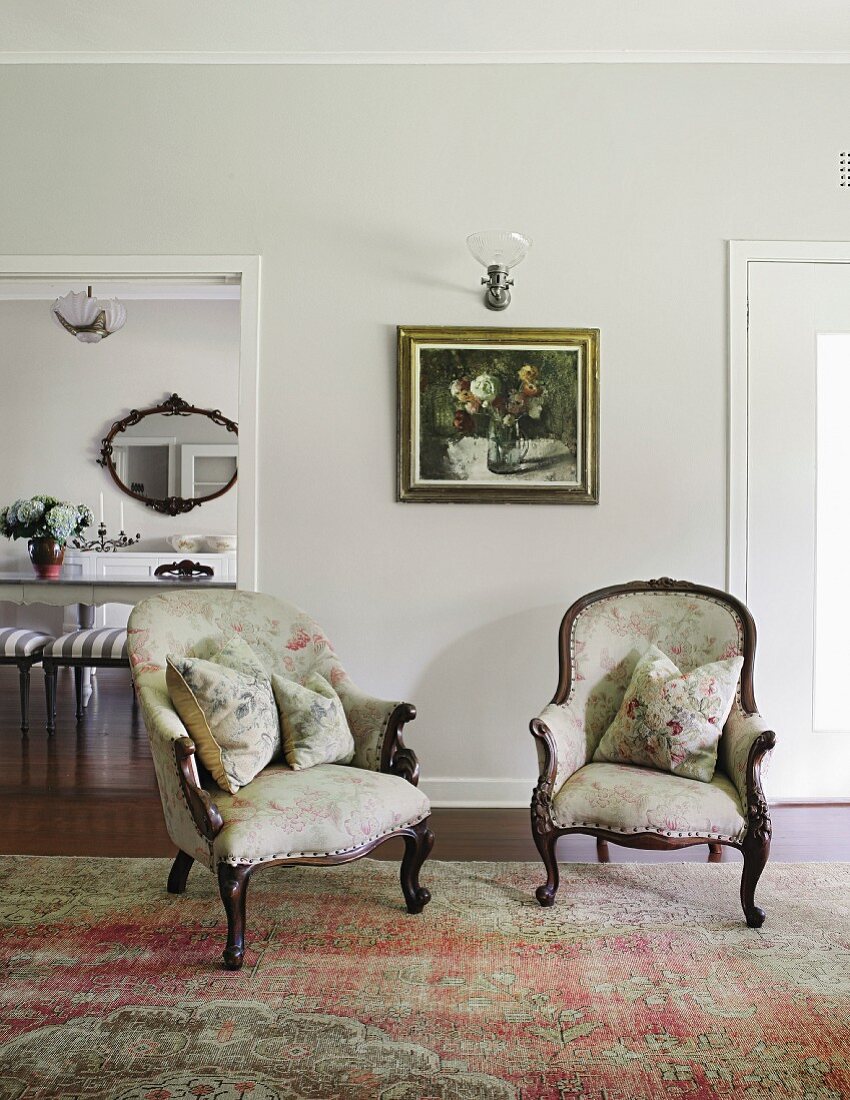 Two antique armchairs with cushions on rug in front of painting and sconce lamp on wall next to open doorway leading to dining room