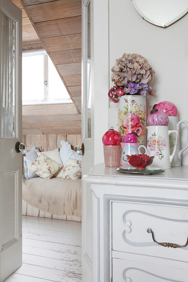 Flowers in vase, ceramic jugs and glass spheres on white chest of drawers