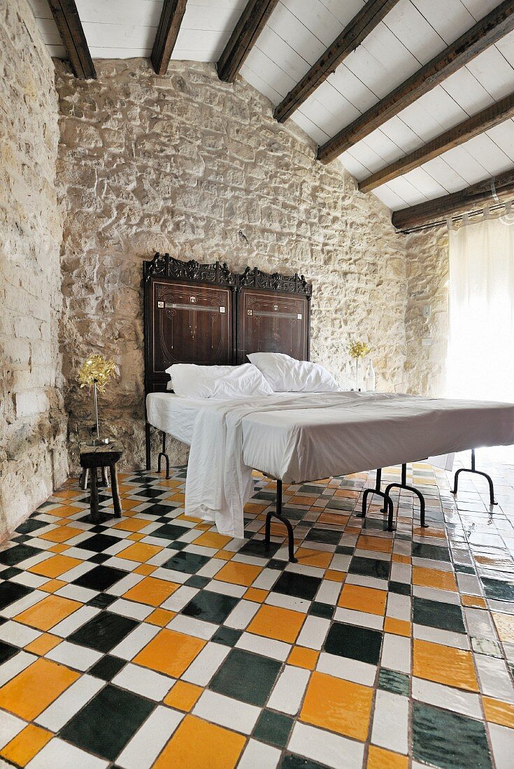 Doppelbett mit antikem Betthaupt vor traditioneller Steinwand, mehrfarbiger Fliesenboden mit geometrischem Muster