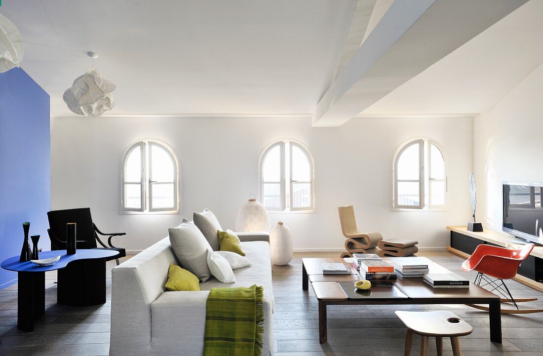 Helles Sofa, Couchtish, Designerstühle und Lowboard im Loungebereich mit Rundbogenfenstern; blauer Raumteiler und Designertisch