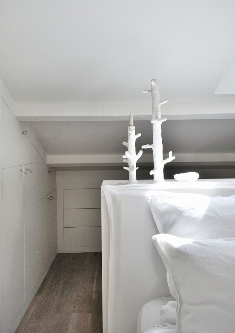 Space-saving fitted cupboards under sloping ceiling and artistic candlesticks in white bedroom