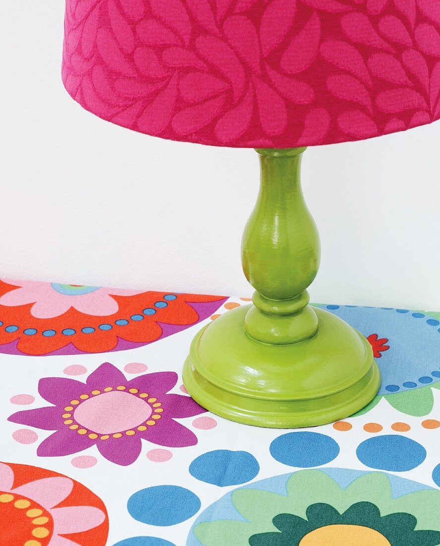 Green-painted table lamp base with pink-patterned lampshade on tablecloth with retro pattern of colourful flowers