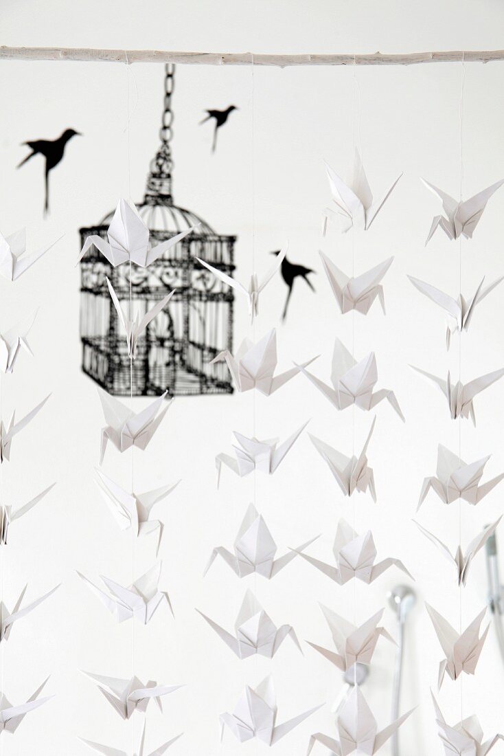 Mobile made from many white origami paper cranes hung from white wooden rod in front of black birdcage motif