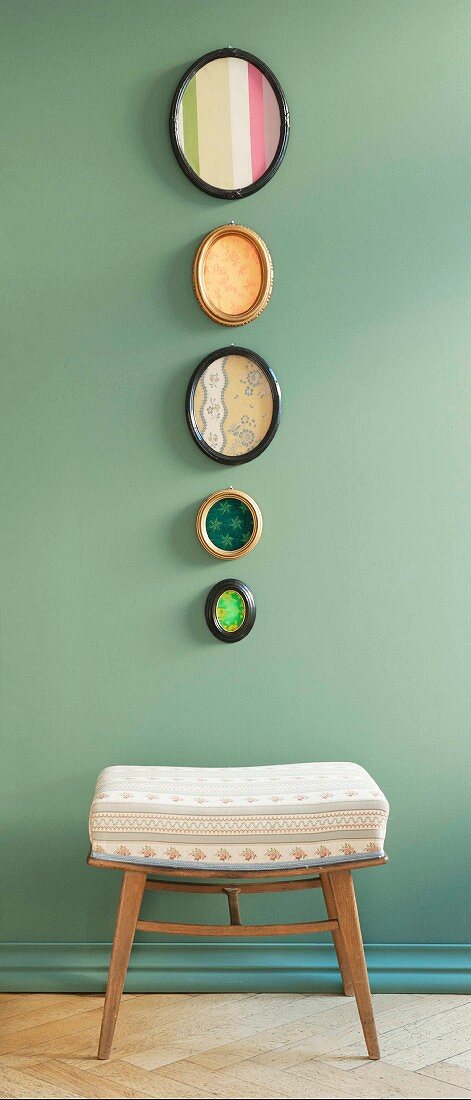 Biedermeier fabric patterns in oval photo frames hanging on a pastel-green wall above an upholstered stool