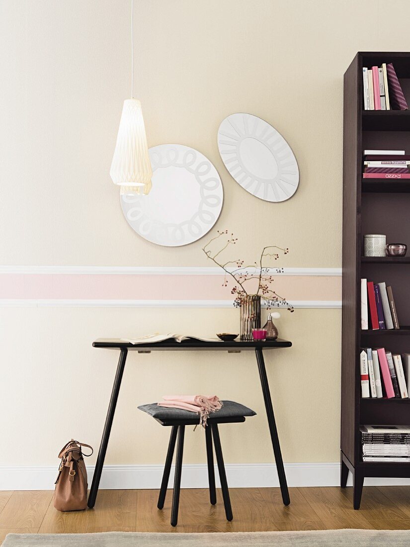 A black table with a stool and a bookcase in front of an elegant wall design with plastered mouldings and pastel-coloured non-woven wallpaper