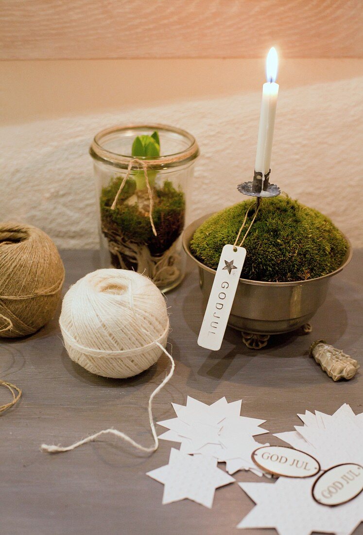 Still-life arrangement of paper stars, ball of string and lit Christmas-tree candle in bowl of moss