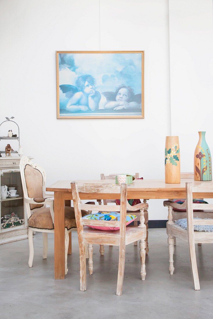 Esstisch mit Vintage Armlehnstühlen auf Betonboden vor gerahmtem Bild mit Engelmotiven