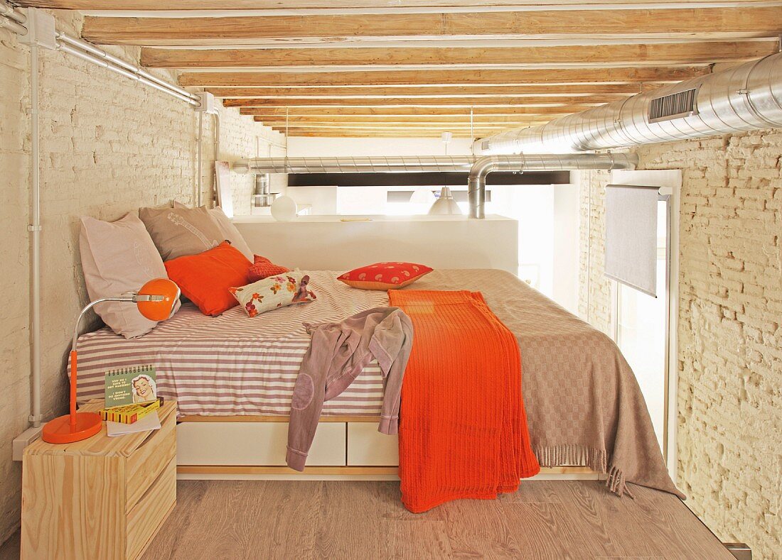 Schlafebene mit orangefarbenen Akzenten in Loftwohnung unter sichtbaren Balken und Lüftungsrohr