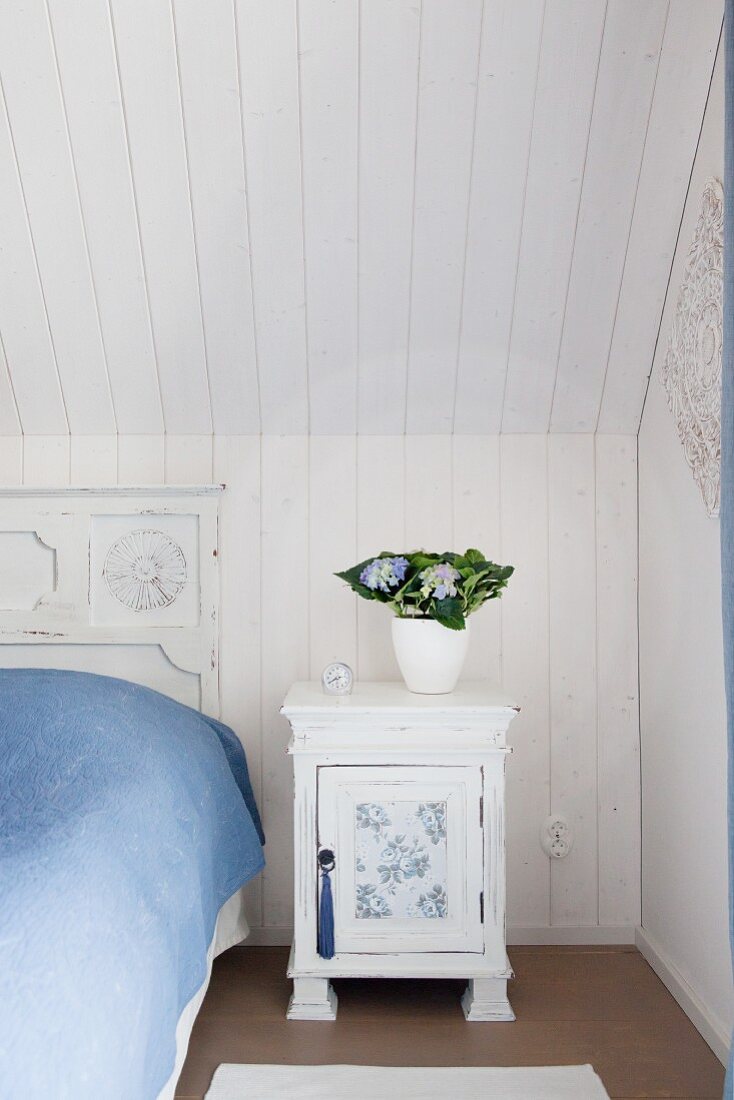 Vintage white bedside cabinet next to bed with vintage headboard below wood-panelled sloping ceiling