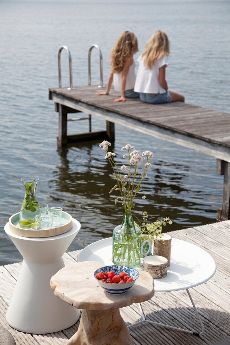 Drinks, flowers and fruit on three side tables next to lake with two girls sat on jetty in background