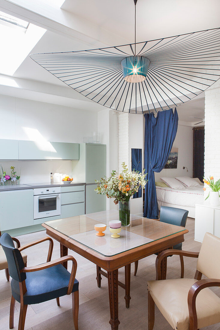 Armchairs around dining table below designer lamp in front of mint-green kitchen counter and curtain screening bedroom