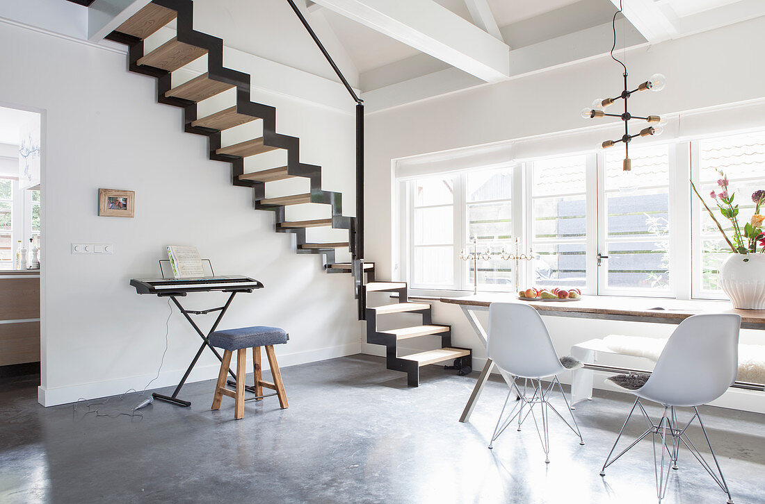 Dining table, classic chair and keyboard below zigzag staircase in open-plan interior with concrete floor