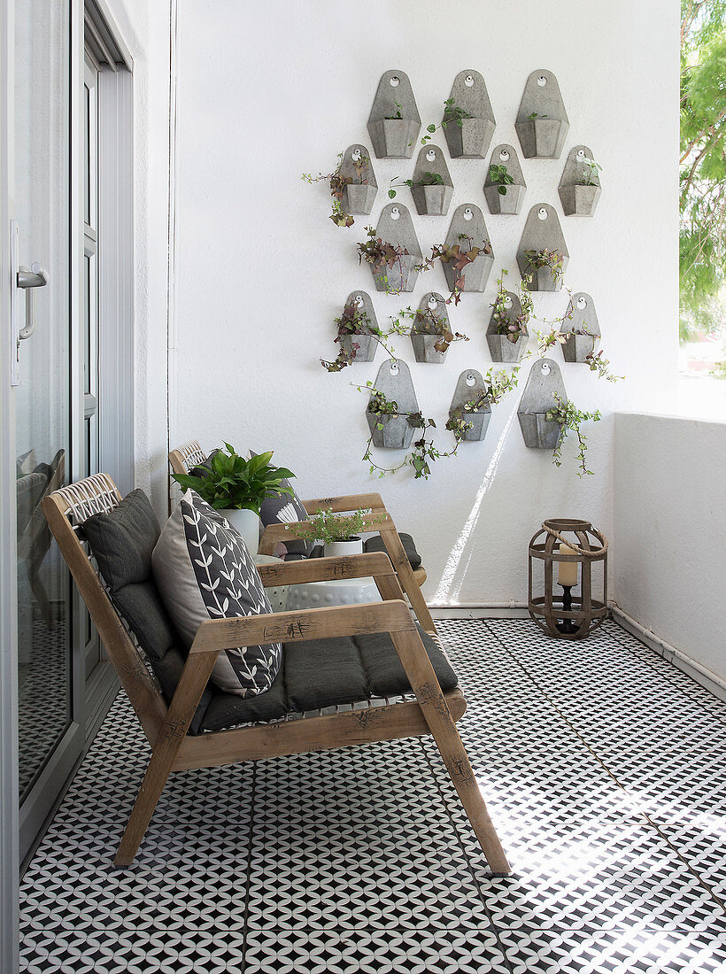 Wooden armchairs with cushions, black and white floor tiles and foliage plants in wall-mounted pots on balcony