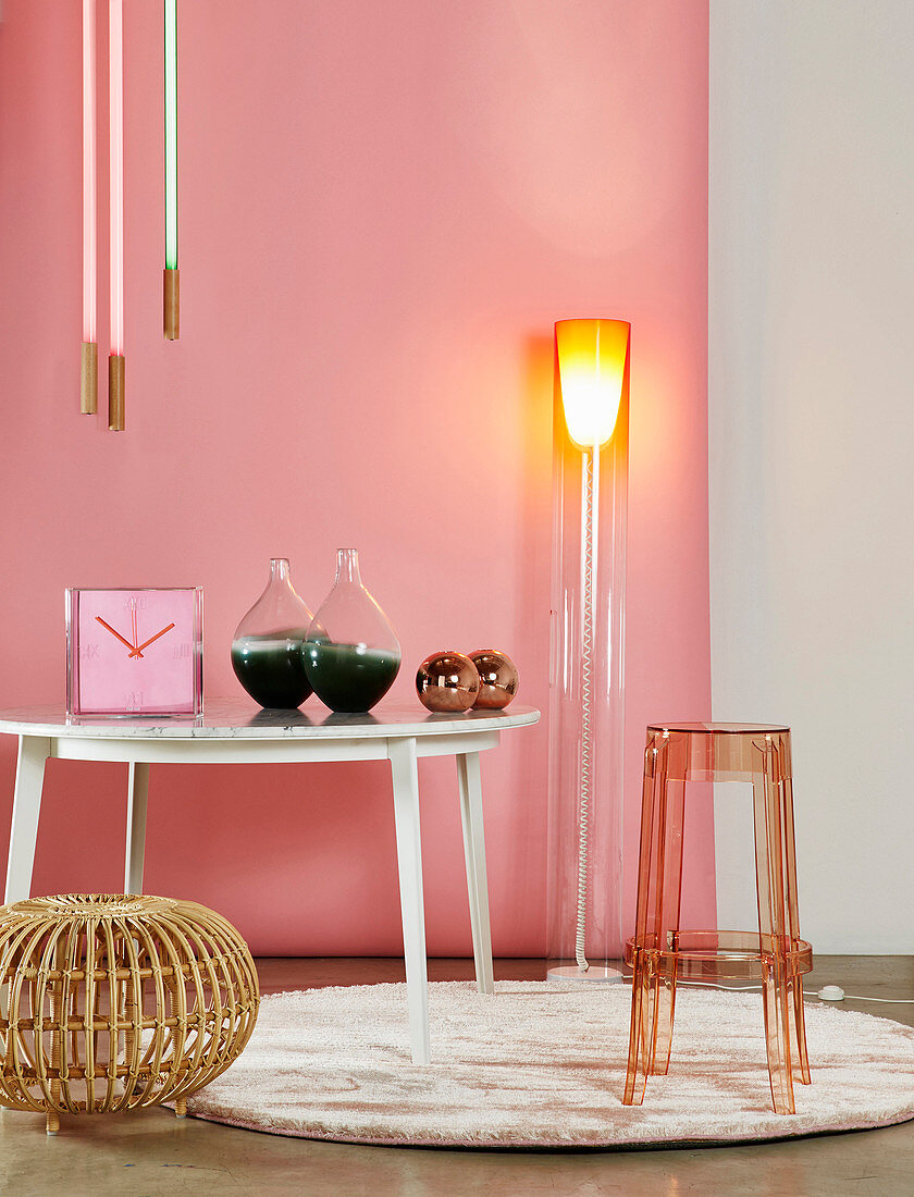 Designer-Stehlampe, Tisch mit Uhr und Glasvasen, transparenter Barhocker und Rattanhocker vor rosa Wand