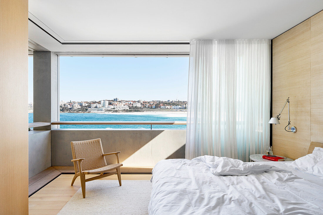 Doppelbett, Wandleuchte und Designer Armlehnstuhl vor Panoramafenster mit Meerblick