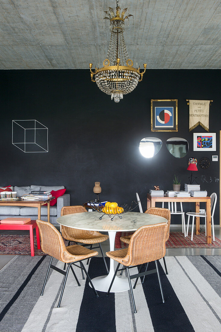 Round table and rattan chairs below chandelier in front of wall painted with chalkboard paint