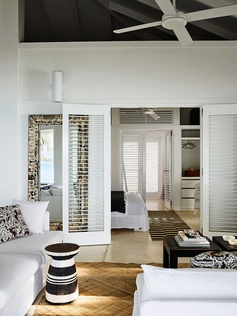 Living room with white upholstered furniture and black and white side table, view through open double doors into the bedroom