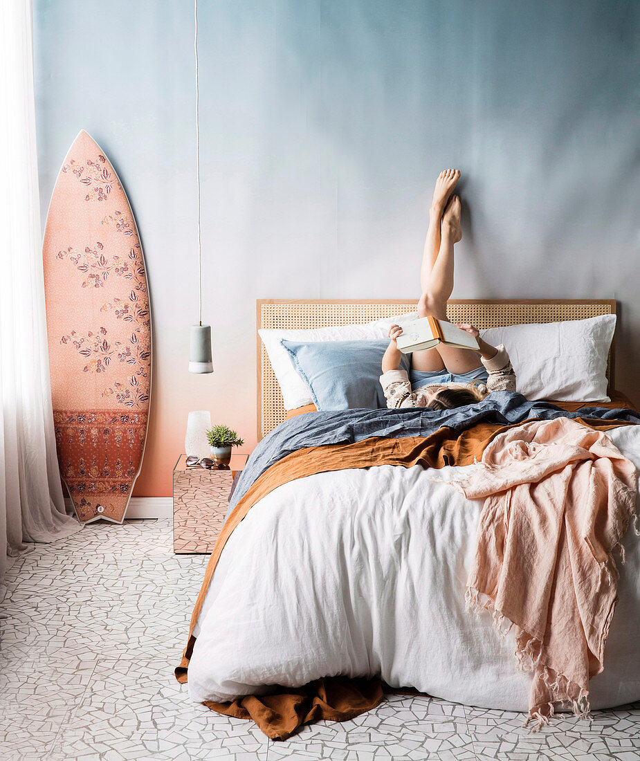 Pastel-colored bedroom, young woman on double bed, pendant lamp and surfboard next to it