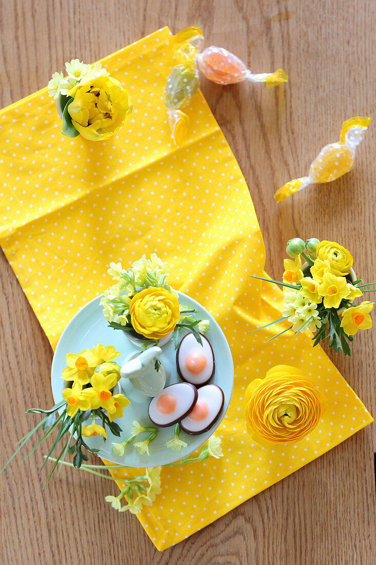 Hasen-Etagere mit Eier-Konfekt und Eierbecher mit Frühlingsblumen auf gelbem Tuch