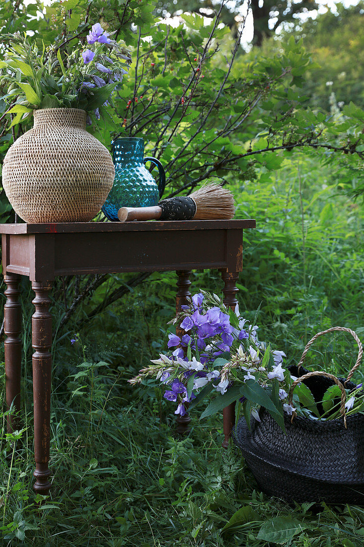 Bastvase und blaue Glaskanne auf Tisch davor Korb mit Blumen im Garten