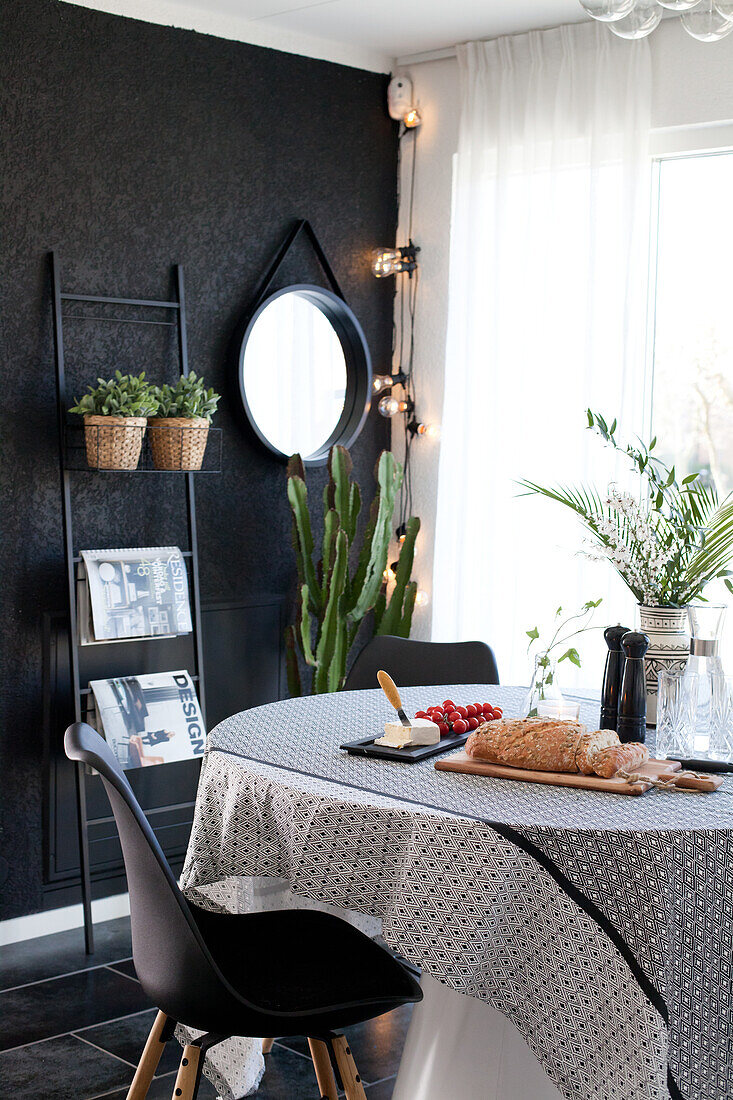 Round table with tablecloth and classic chairs in front of potted plants and magazines on ladder shelves and round mirror