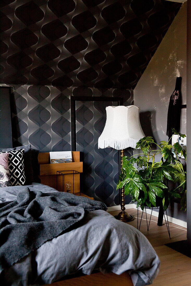 Double bed with dark bed linen, wooden bedside cabinet, standard lamp and houseplant in bedroom with black wallpaper