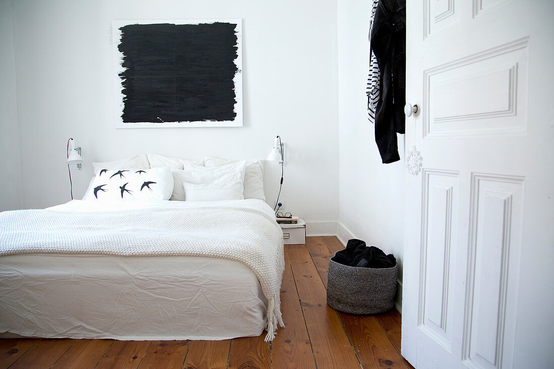 Doppelbett mit weißer Decke, darüber schwarzes Bild im Schlafzimmer mit Holzdielenboden