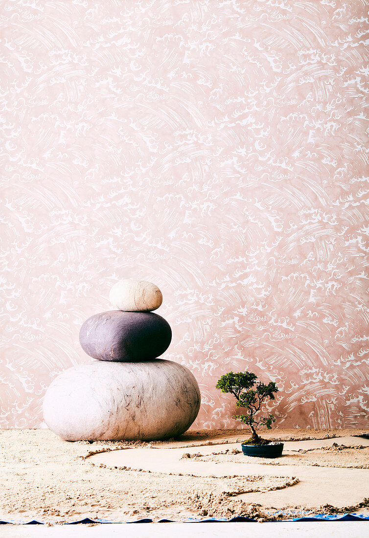 Kieselsteine aus Filz und ein Bonsai auf Sand vor rosafarbener Tapete