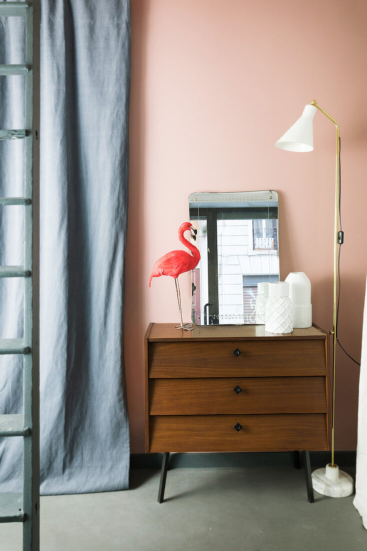 Standard lamp next to vases, mirror and flamingo ornament on top of chest of drawers against pink wall