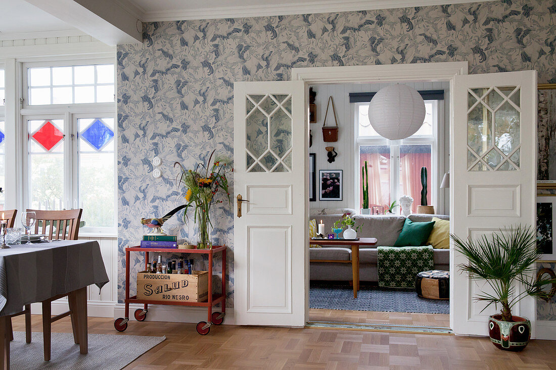 Open double doors painted white, vintage serving trolley and house plants against patterned wallpaper