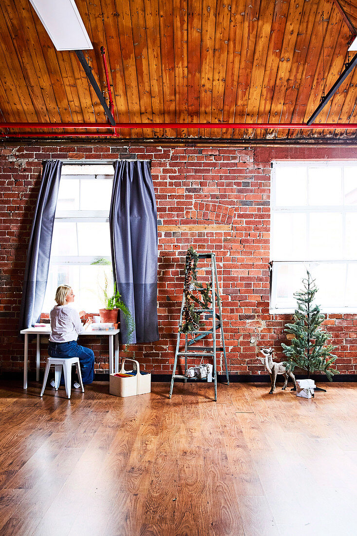 Frau am Tisch vor Fenster, daneben Leiter mit Weihnachtsdekoration, Rehfigur und kleiner Tannenbaum vor Backsteinwand