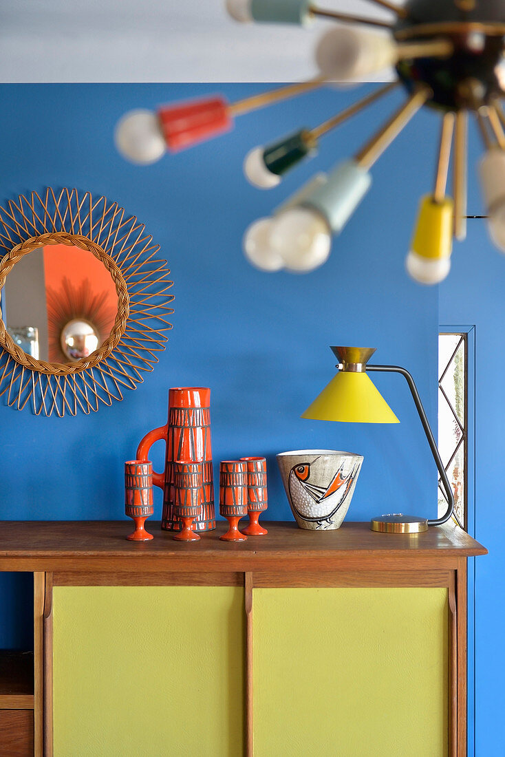 Keramiksammlung und Tischlampe auf Retro Sideboard, darüber Sonnenspiegel an blauer Wand